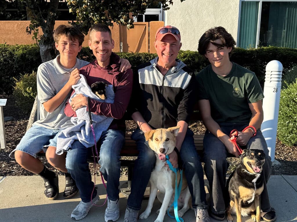 A group of four people sitting on a bench outside, smiling. Two men hold leashed dogs while two boys, one cradling a small dog wrapped in a blanket, sit beside them. The scene reflects a joyful moment, emphasizing the theme of adoption.