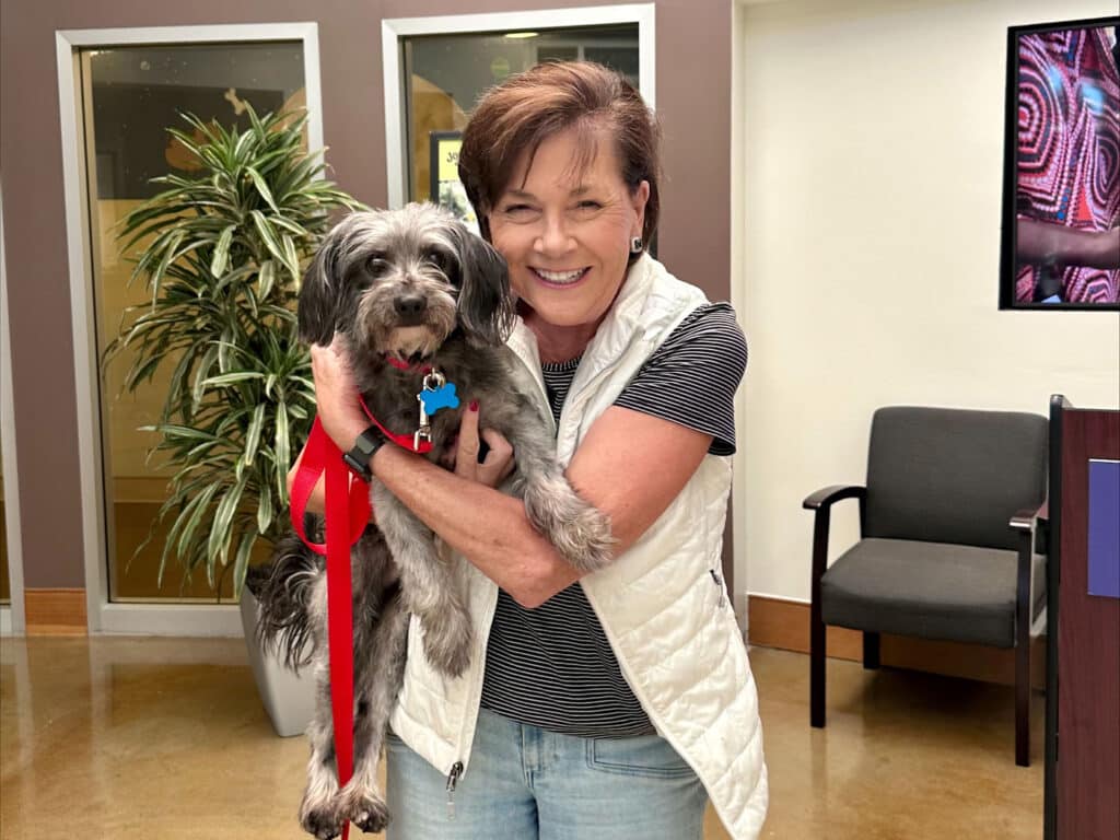 A woman holding a small gray dog with a red harness in a welcoming indoor space, surrounded by plants. The woman is smiling as she poses for the camera, emphasizing the theme of pet adoption.