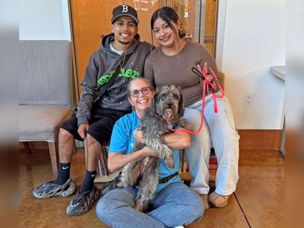 A young man and woman sit on a bench alongside a woman holding a small dog. They all smile at the camera in a brightly lit room. The image highlights a joyful moment related to adoptions at Joybound.