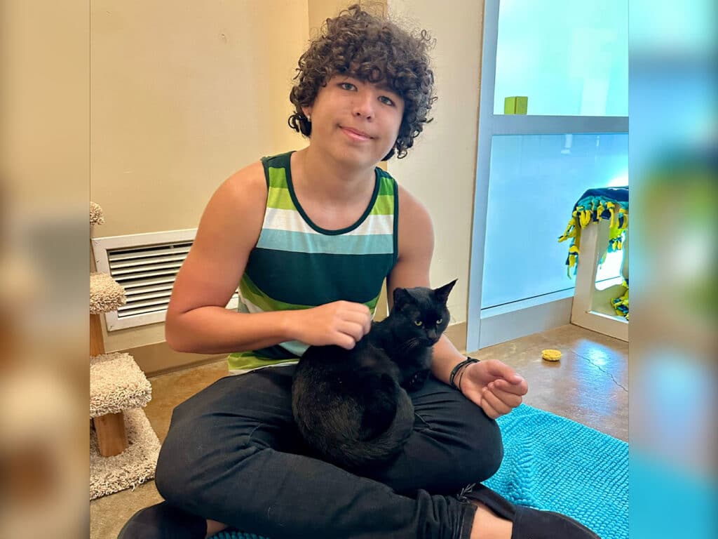 A young boy with curly brown hair wearing a striped tank top sits cross-legged on a blue rug in a bright room, gently holding a black cat in his lap. The setting includes a scratching post and colorful toys, supporting the theme of pet adoption.