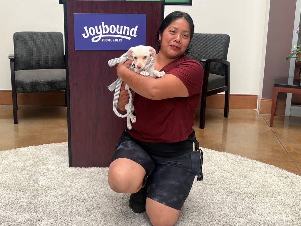 A woman wearing a red shirt and black shorts kneels on a gray rug, holding a small white puppy. Behind her, a sign with the logo "Joybound People & Pets" is visible. The setting appears to be a pet adoption center.