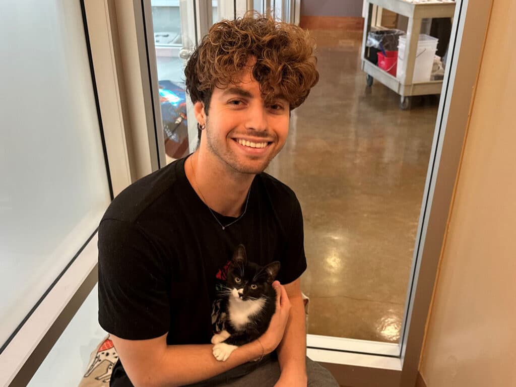 A young man with curly hair in a black shirt is sitting on the floor, holding a small black and white kitten in his lap. The setting is a well-lit adoption space with a clean, polished floor and shelves in the background. This image relates to Adoptions at Joybound.