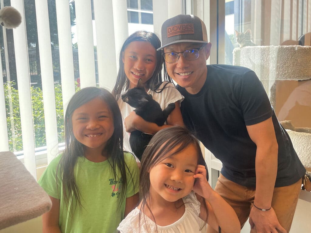 A man wearing a cap stands with three smiling girls indoors. The man holds a black kitten. Sunlight streams through large windows, illuminating the cozy space adorned with cat trees. The image reflects a joyful moment associated with pet adoption.