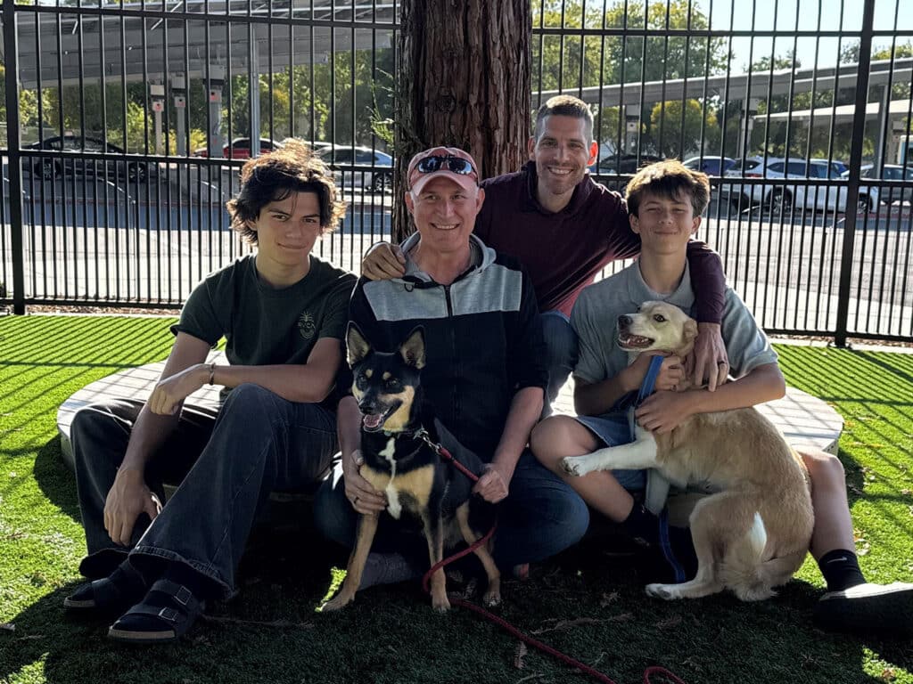 A group of four people, including two teens and two adults, smile while seated on grass near a tree. They are accompanied by two dogs, one being held by a person and the other sitting by their side, emphasizing a positive adoption and bonding experience.