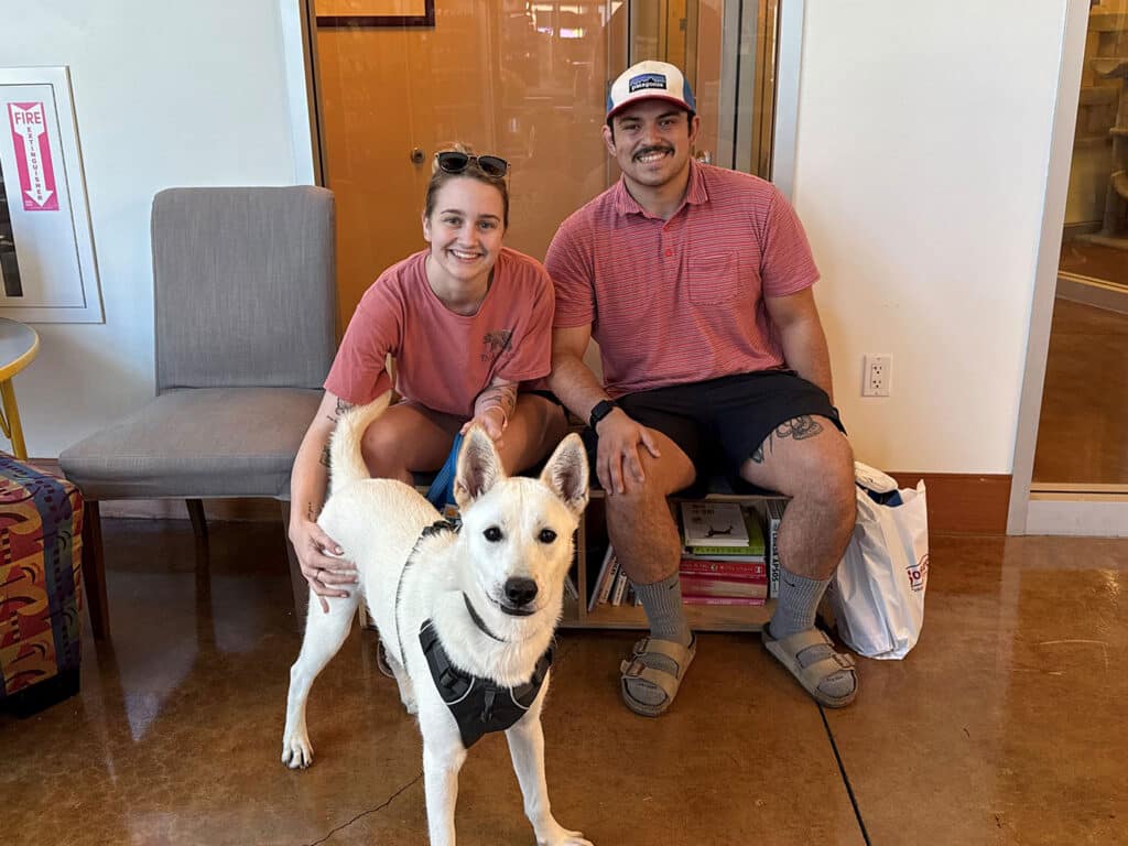 A woman and a man sit together on a bench in a pet adoption center, smiling at the camera. In front of them is a white dog wearing a harness. The setting appears welcoming, with a cozy interior and a stack of books in the background, highlighting Adoptions at Joybound.