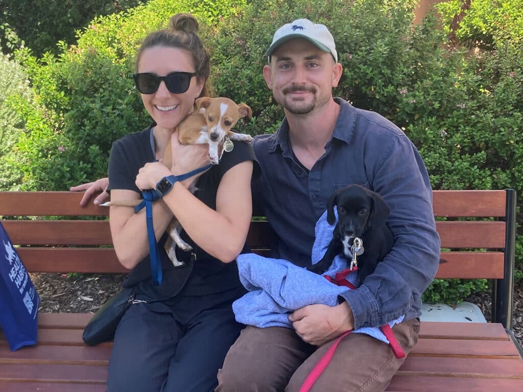A woman and a man sit on a bench, each holding a small dog. The woman, wearing sunglasses, holds a tan and white puppy, while the man cradles a black puppy wrapped in a blue cloth. Green bushes are visible in the background, suggesting a park setting. This image relates to pet adoption.