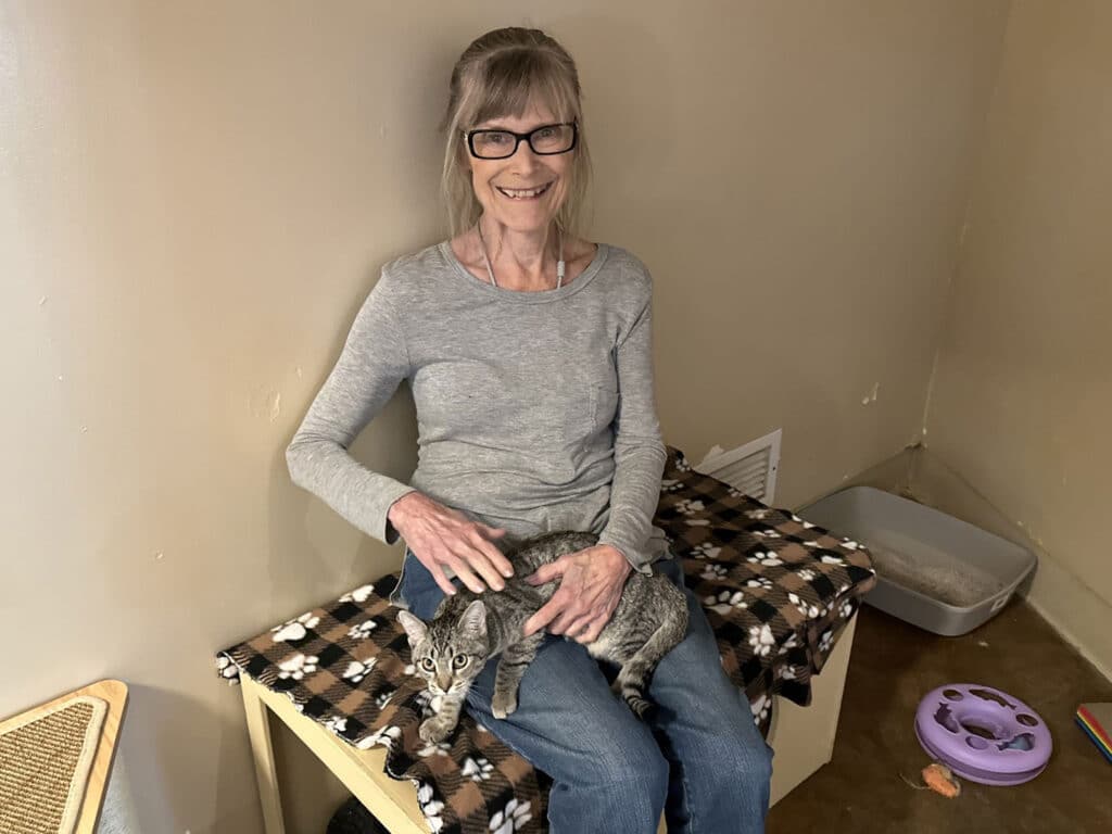 A woman sitting on a bench in a room, holding a gray tabby cat on her lap. She has long hair and glasses, smiling softly. In the background, there is a litter box and a purple cat toy. The setting suggests a space related to pet adoption.