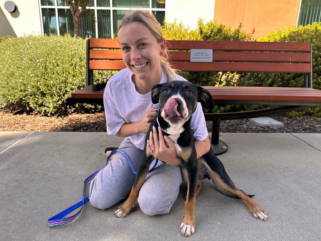 A woman in casual attire kneels beside a black and tan dog, both smiling in a sunny outdoor setting. They sit on a paved surface near a bench with greenery in the background. The image highlights the joyful moment of adoption.