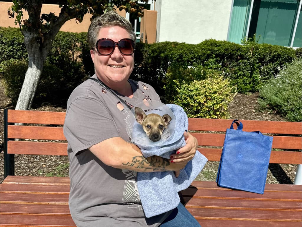 A woman wearing sunglasses sits on a bench, smiling while holding a small dog wrapped in a blanket. A blue bag is placed beside her, with greenery and a building visible in the background. This image relates to adoption themes.