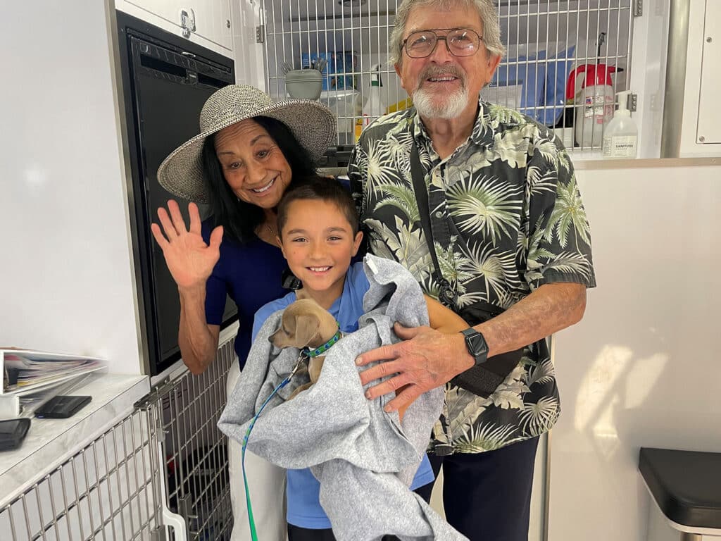 A young boy holding a small dog wrapped in a blanket stands between an elderly couple in a pet adoption setting. The woman waves, wearing a wide-brimmed hat, while the man smiles beside her, dressed in a floral shirt. The background displays a pet care environment.