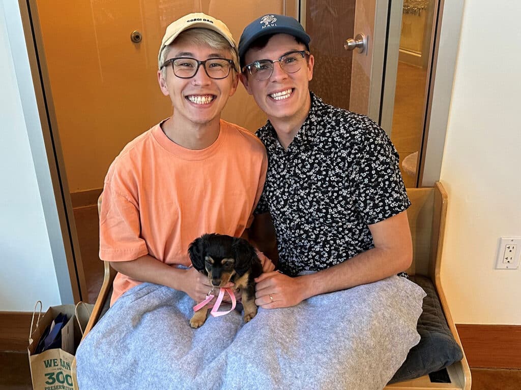 Two smiling men sitting together on a bench, holding a small puppy with a pink collar. The men are wearing casual clothing, with one in a light orange t-shirt and the other in a black patterned shirt. The background shows a modern indoor space.