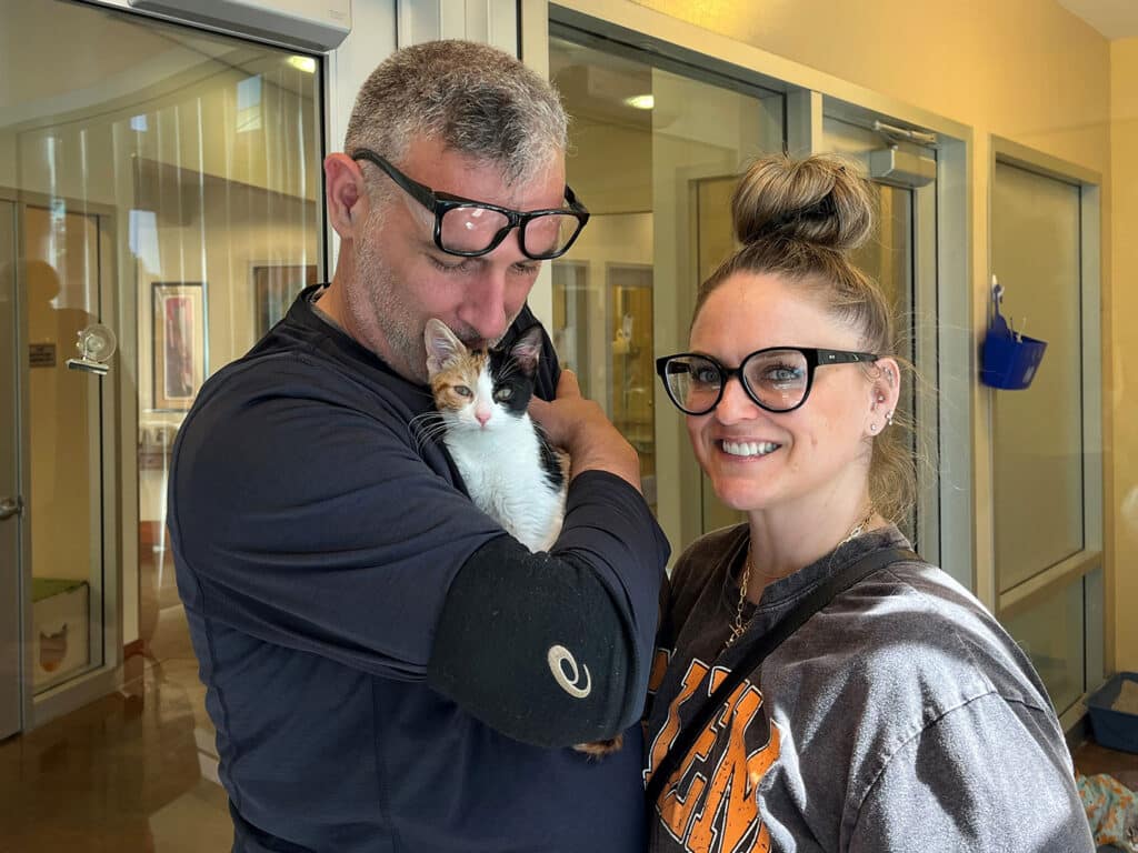 A man wearing glasses holds a small calico kitten in his arms while standing next to a woman with glasses. They both smile, standing in a bright room, possibly in an animal adoption center, highlighting the theme of adoption.