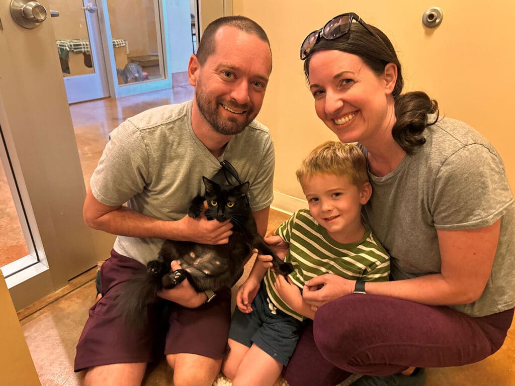 A smiling family of three, including a man holding a black cat, a woman sitting next to him, and a young boy wearing a striped shirt, all sitting on a floor in a bright room. This image relates to Adoptions at Joybound.