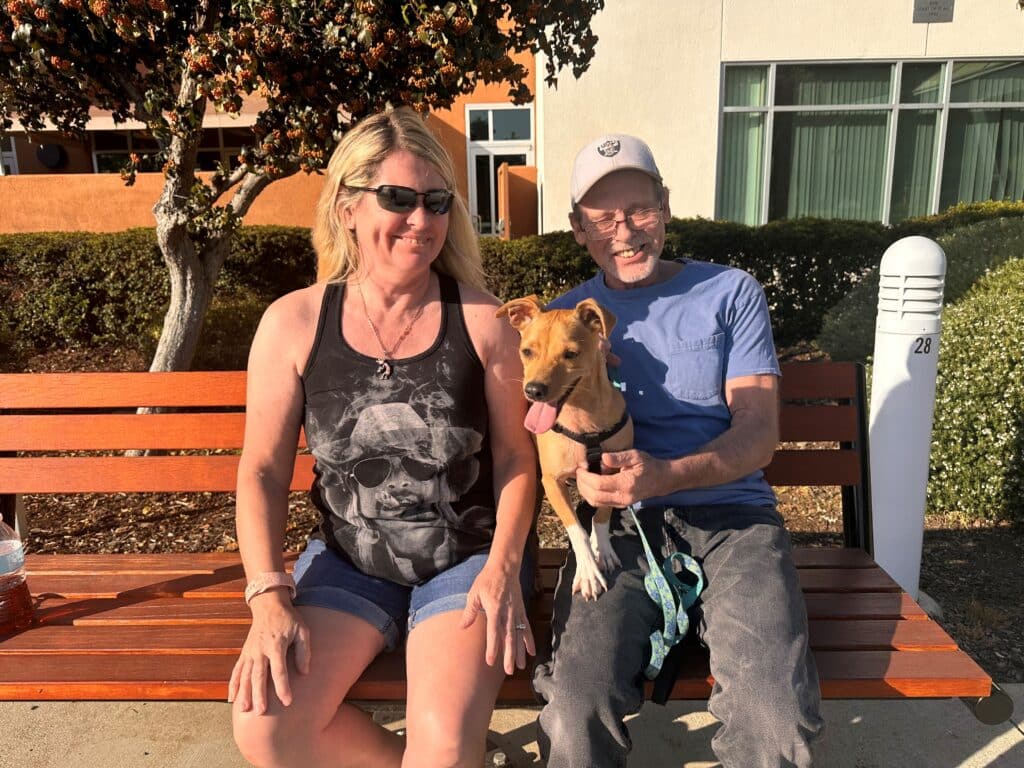A man and a woman sit together on a wooden bench outside. The woman wears sunglasses and a black tank top, and the man has a cap and a blue shirt. They hold a small dog between them, smiling in a sunny outdoor setting.