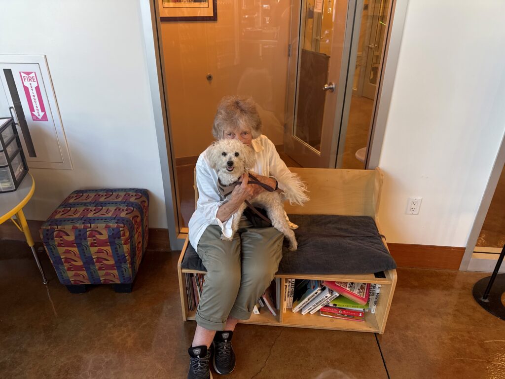 An elderly woman seated on a wooden bench, holding a small white dog in her arms. The background features a bookshelf and a window, with a bright, open atmosphere in the room. A patterned ottoman is visible to the left.