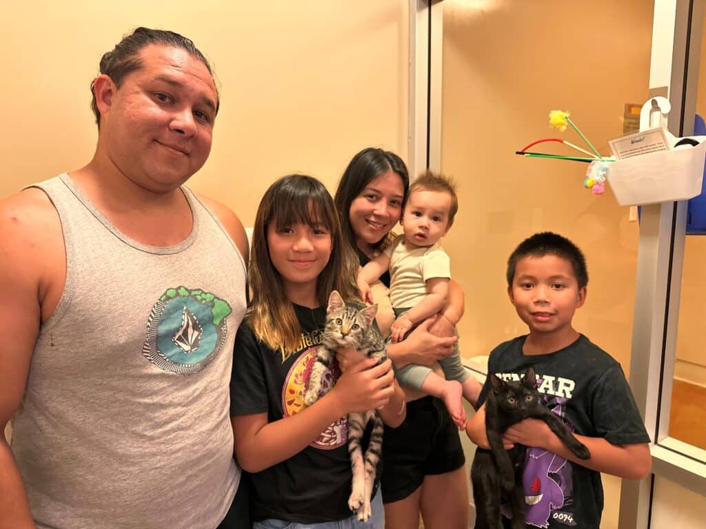 A family of five poses for a photo indoors. The father stands on the left wearing a gray tank top. A girl holds a tabby cat, while another girl with long hair smiles. A baby is held by the mother, and a boy stands on the right with a black cat.