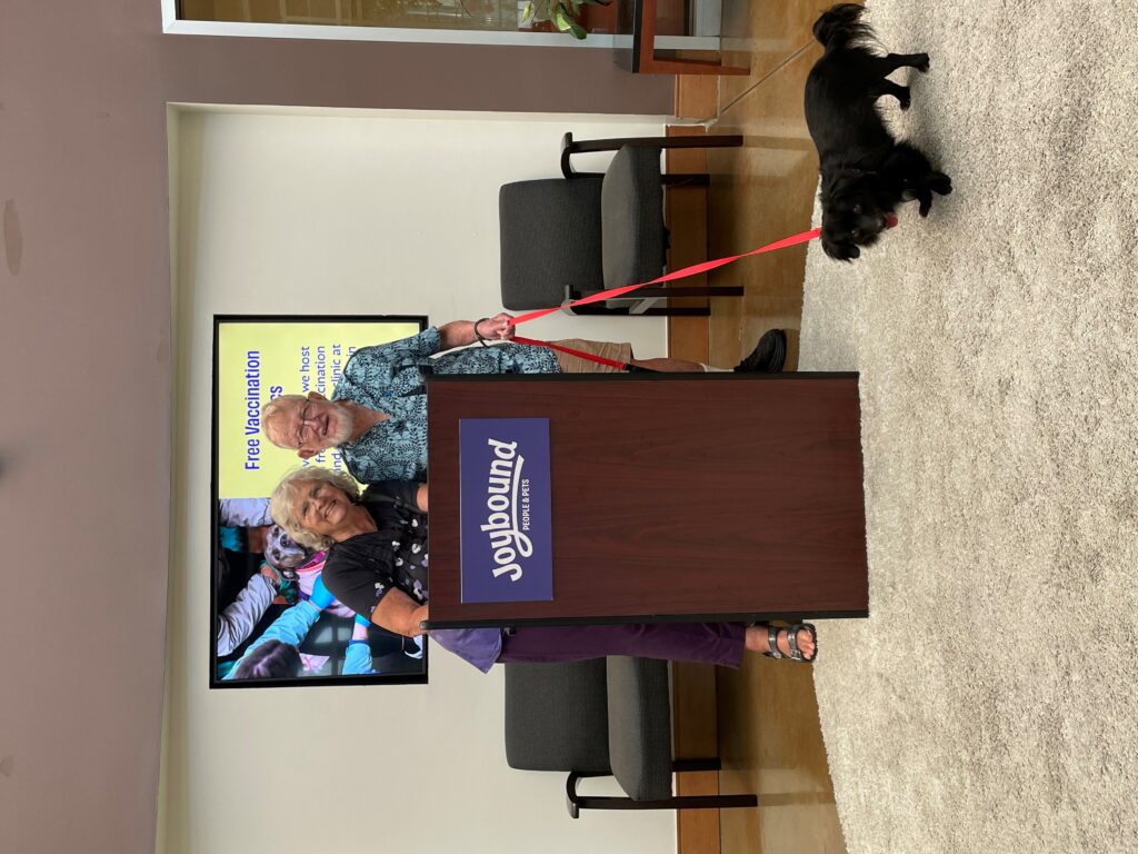 Two individuals stand behind a podium with the "Joybound" logo, smiling at the camera. A small black dog is on a leash near them. In the background, there is a screen displaying text related to "Free Vaccination." The setting appears to be a casual event space with soft furnishings.