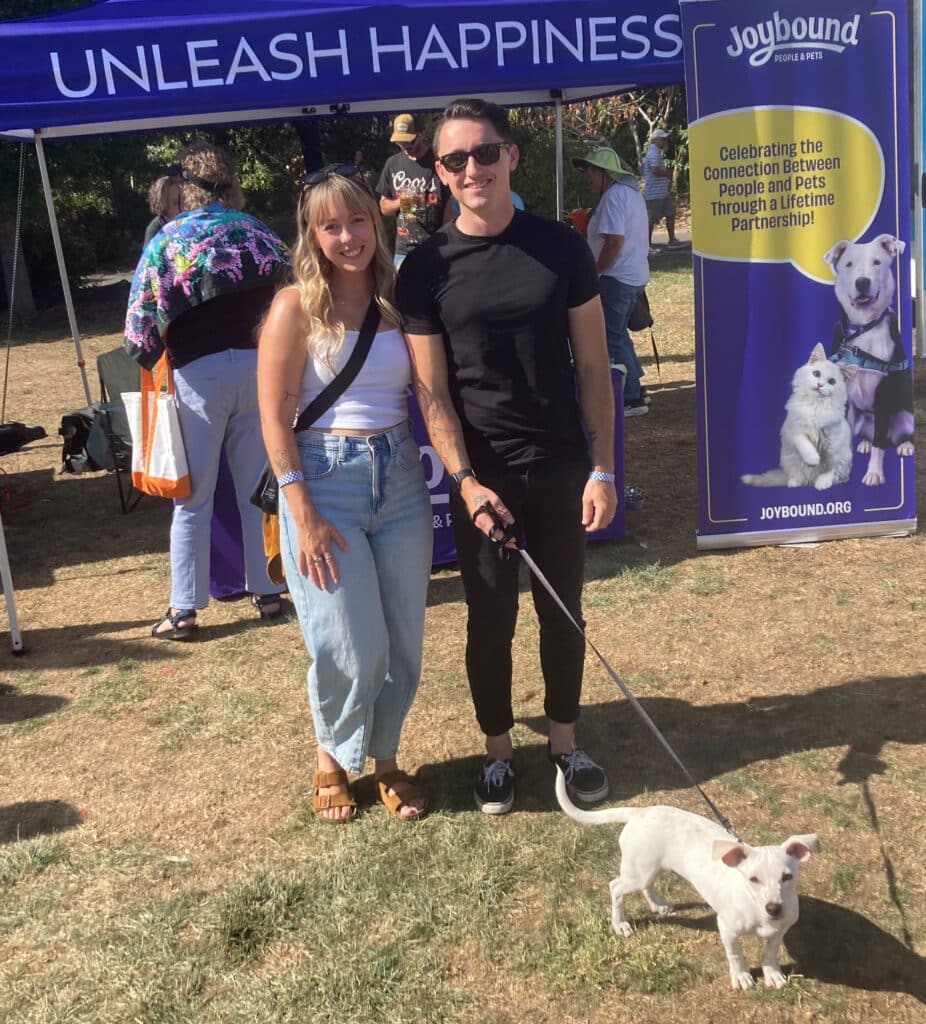 A man and woman stand smiling in front of a Joybound booth at an outdoor event. The woman has long hair and wears a light shirt with jeans, while the man wears a black shirt and sunglasses. They are holding the leash of a small white dog.