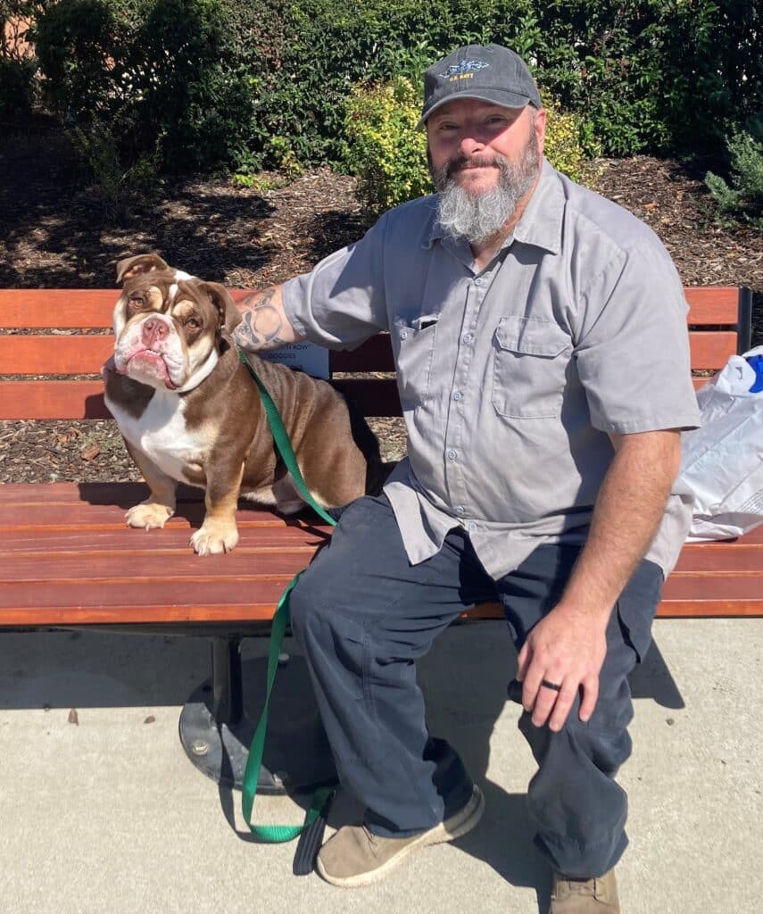 A man sitting on a park bench with a brown and white bulldog beside him. The man is wearing a gray shirt and a cap. The background features greenery and shrubs, indicating an outdoor setting.