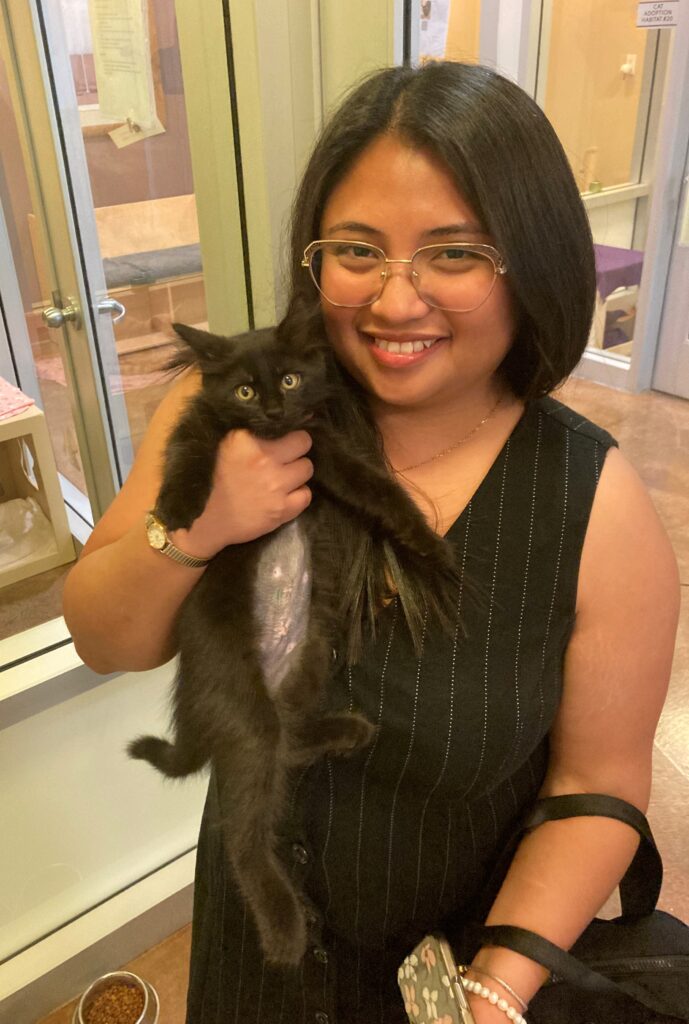 A woman holding a black kitten in a pet adoption center, smiling. She wears glasses and a black striped outfit. The background shows glass enclosures with other animals, indicating a warm and inviting atmosphere.