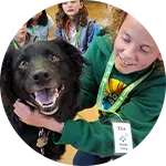A child with curly hair smiles while holding a black dog, both positioned in a cheerful indoor setting. Other children are seated in the background. This image highlights programs focused on rescuing animals in need.