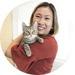 A woman with shoulder-length hair is smiling while holding a gray tabby cat against her chest. She is wearing a red top and standing in a bright, softly lit indoor space. The image supports topics related to animal care and training.