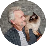 An older man with gray hair smiles while holding a Siamese cat on his shoulder. The background is softly blurred, highlighting the man and the cat. The image conveys a sense of care, relevant to animal rescue donations.