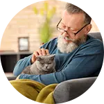 An older man with glasses is sitting comfortably in a chair, gently petting a gray cat on his lap. The setting is cozy, with a soft blanket and indoor plants visible in the background, highlighting the theme of animal rescue donations.