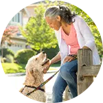 An older woman sitting on a wooden fence interacts with a tan dog on a leash. The background features a residential neighborhood with greenery. This image illustrates the importance of animal rescue donations.