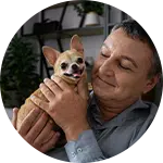 A man holding a small tan Chihuahua with a happy expression, sitting in a cozy indoor setting. Shelves with plants can be seen in the background. The image emphasizes the bond between the man and the dog, relating to animal rescue donations.