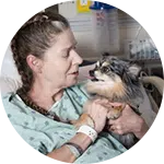 A woman in a hospital bed interacts with a small dog, smiling at each other. The setting is a medical facility, with hospital equipment visible in the background, highlighting efforts toward rescuing animals in need.