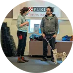 A woman and a man stand in a training space, discussing dog training techniques. The man holds a small dog on a leash. The background features equipment and a banner promoting dog training services.