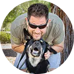 A man wearing sunglasses is joyfully smiling while holding a black dog close to his face. The background features greenery, indicating an outdoor setting. The image depicts a moment of connection, emphasizing efforts in rescuing animals in need.