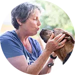 A woman with short gray hair gently kissing a brown dog, showing affection. They are outside, with natural light illuminating the setting, highlighting their connection and the theme of rescuing animals in need.