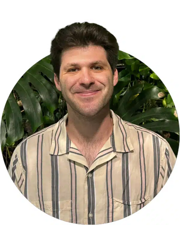 A man with short dark hair smiles while wearing a striped shirt, set against a background of green plants. This image could represent dog training workshops or similar training programs.