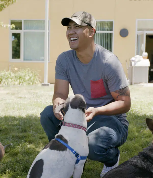 A smiling man in a gray t-shirt and baseball cap sits on grass with a small dog at his feet. The background features a yellow building and greenery, suggesting a relaxed outdoor setting. This image connects to the topic of veterans benefits through companionship.