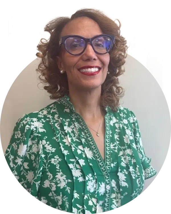 A woman with curly hair wearing blue glasses and a green floral blouse smiles for the camera. The image is circular, highlighting her features. This photo is associated with the Joybound Board of Directors.