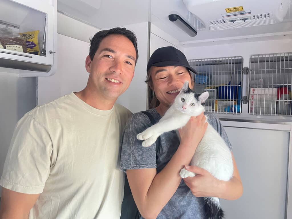 A man and a woman stand together in a bright space, smiling for the camera. The woman holds a white cat with black markings. Behind them is a white storage unit with shelves, likely containing food or supplies.