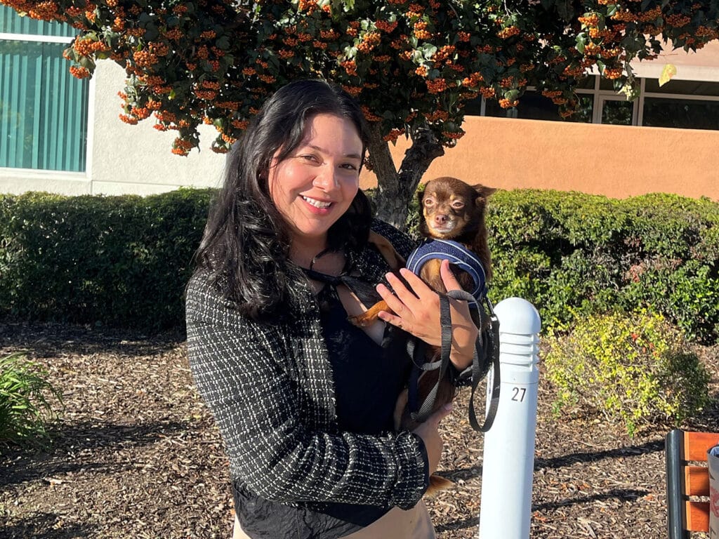 A woman with long black hair smiles while holding a small brown Chihuahua against her chest. They are standing outside near a bush with orange berries and a building in the background. The woman is wearing a black sweater and light pants.