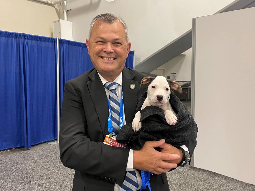 A man in a dark suit, smiling while holding a small puppy wrapped in a black blanket. The background features blue curtains and a neutral-colored wall, indicating an event setting.