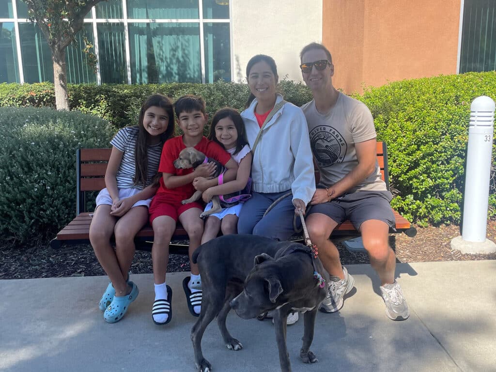 A family of five sits together on a bench in an outdoor setting. Three children, two girls and one boy, smile while holding a small puppy. An adult woman in a light jacket and a man beside her, wearing a T-shirt, relax with a black dog at their feet.