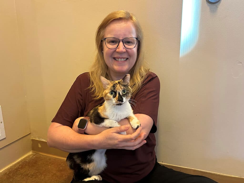 A woman with glasses sits on the floor, holding a calico cat in her arms. She has long hair and is wearing a maroon shirt. The background features a light-colored wall and a doorway, creating a cozy indoor setting.