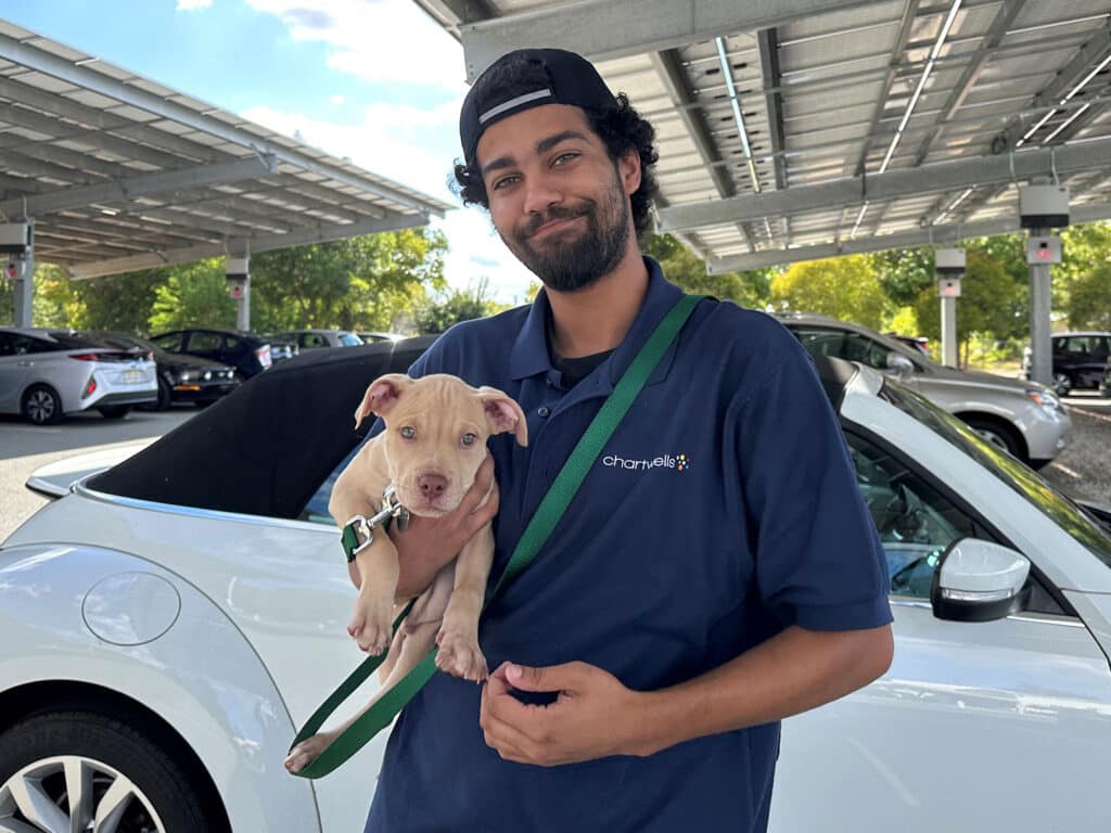 A man wearing a dark blue polo shirt holds a light-colored puppy in his arms, standing next to a white car in a shaded outdoor parking area. Surrounding trees are visible in the background.
