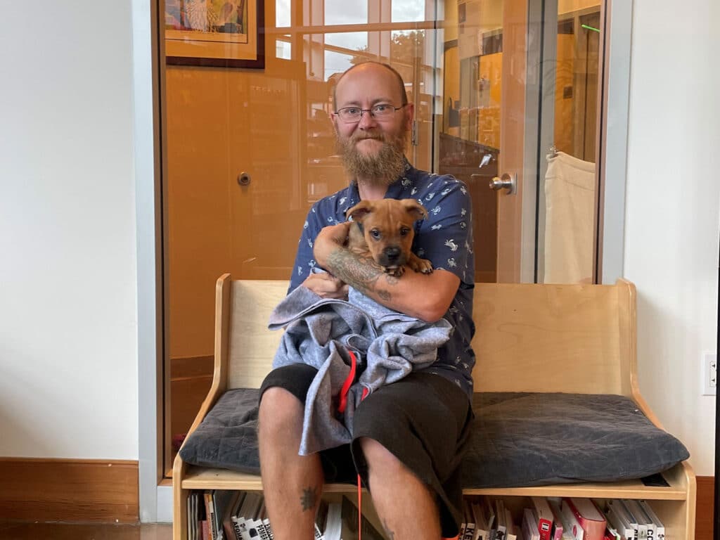 A man with a beard wearing glasses sits on a bench inside a building, holding a small dog wrapped in a blanket. The background features large windows and framed pictures on the wall.