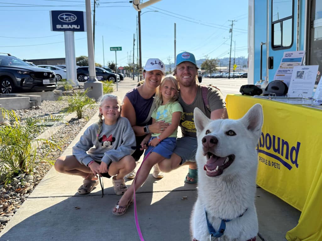 A family of four and a white dog posing together on a sidewalk. Two girls sit in front, one wearing a gray sweater, while another girl stands beside their mother, who is holding the dog on a leash. A man stands behind them. A Subaru dealership and a banner are visible in the background.