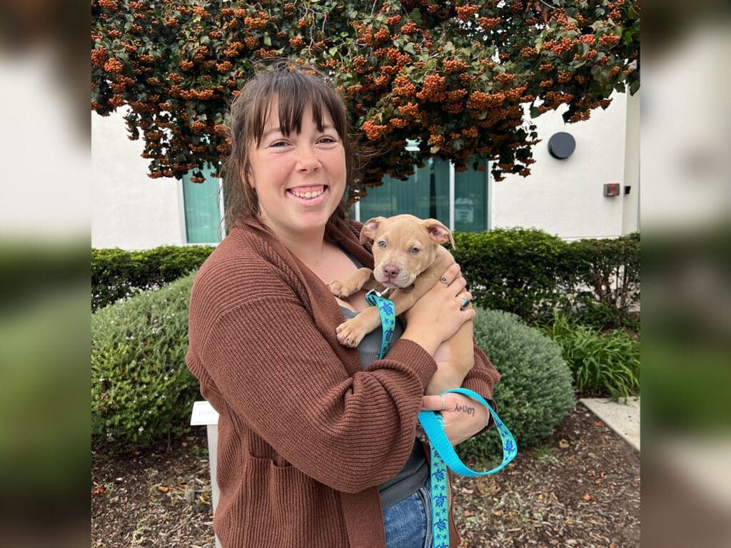 A woman in a brown cardigan smiles while holding a light brown puppy with blue leash in her arms. She stands outdoors in front of a hedge and a tree with orange berries, with a building visible in the background.
