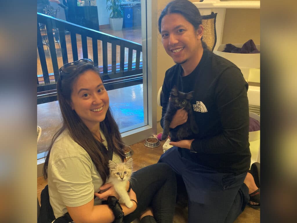 A woman and a man sitting on the floor in a pet adoption center, each holding a kitten. The woman has a light-colored kitten on her lap and is smiling, while the man holds a dark-colored kitten. In the background, there are pet supplies and a window.