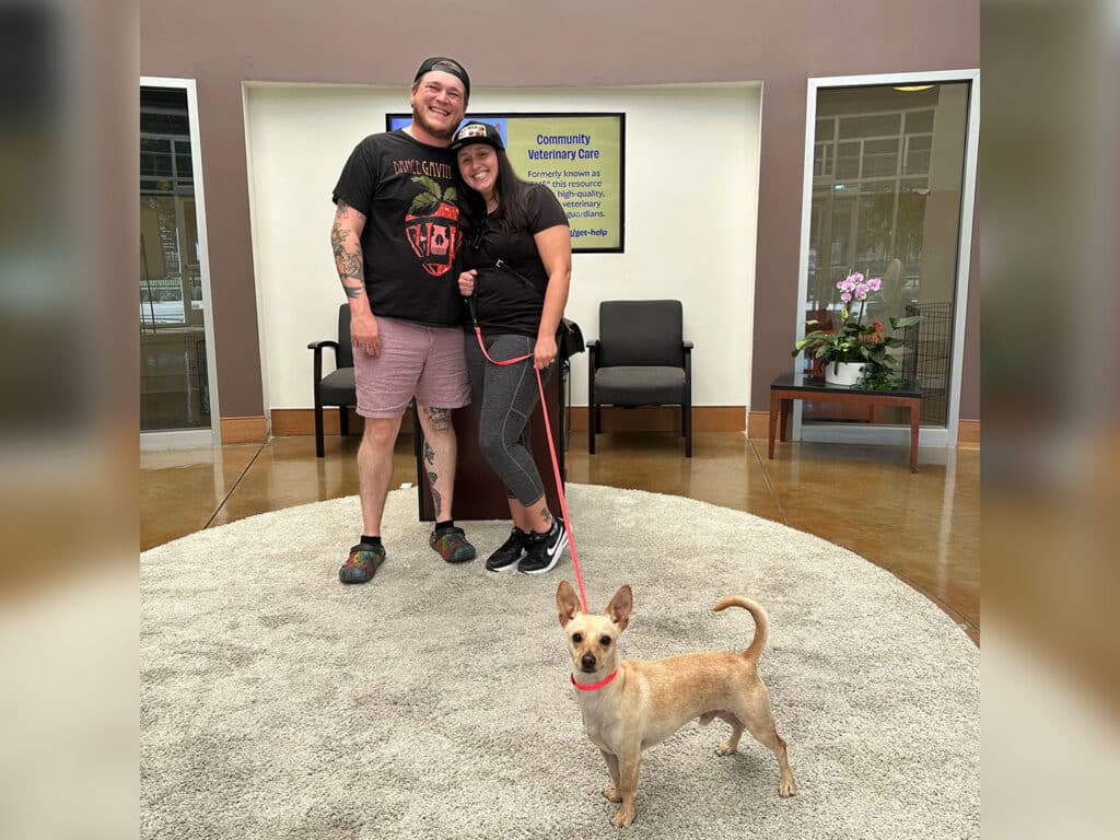 A man and woman pose together in a veterinary clinic with a small dog on a leash. The man wears a black T-shirt and shorts, while the woman wears a black shirt. The clinic’s interior features a waiting area with chairs and a flower arrangement.