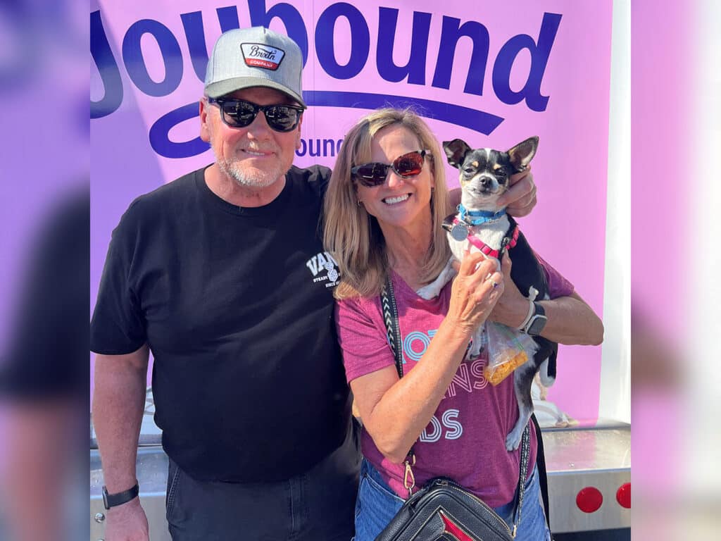 A man and a woman pose together in front of a purple backdrop with the word 'Joybound.' The man is wearing a black shirt and a gray cap with sunglasses. The woman has long hair, wears a maroon shirt, and holds a small dog.