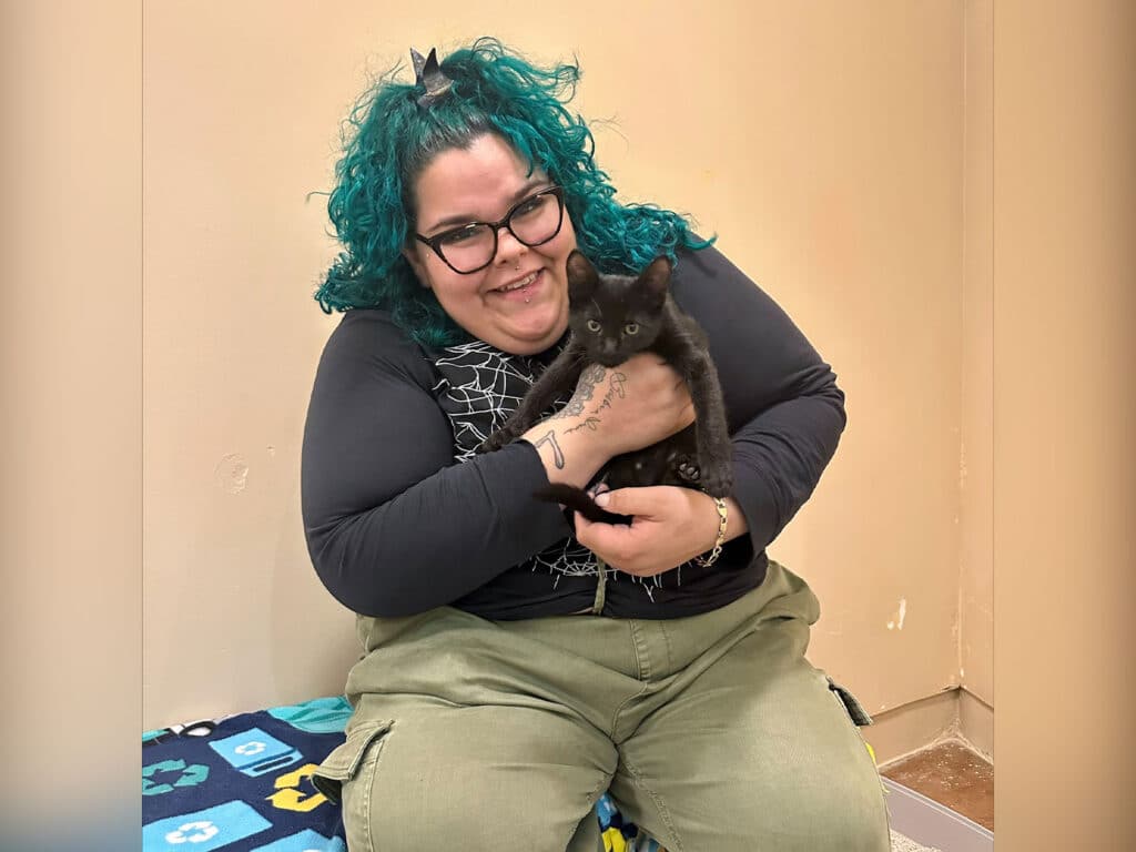 A person with teal curly hair and glasses sits on a blue and green patterned blanket, holding a black kitten. They are smiling, dressed in a dark top and olive green pants, with a neutral wall in the background.